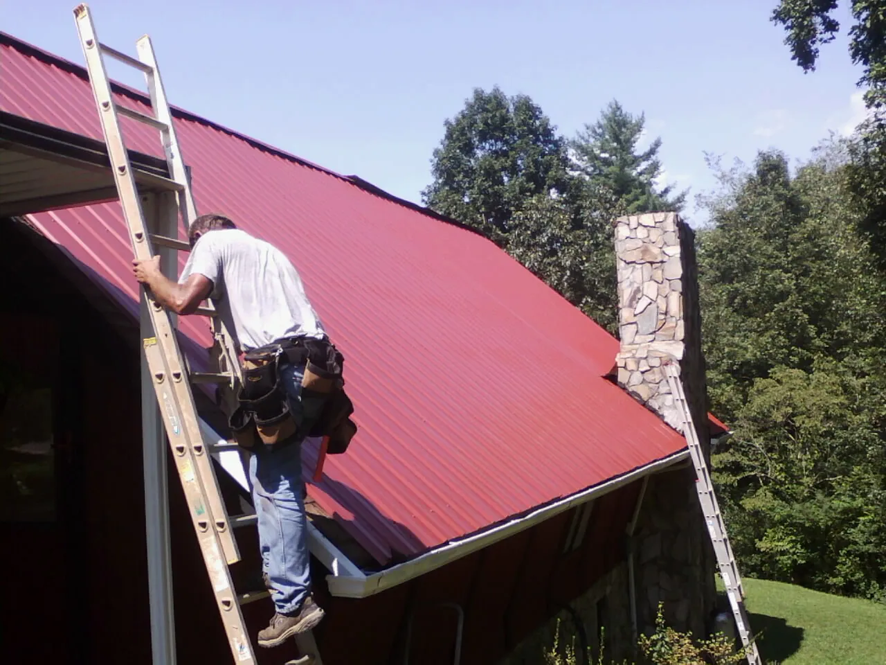 Roofer on a ladder beside a red metal roof and stone chimney — Soffit & Fascia Repair by Roofing Brothers in Orviston